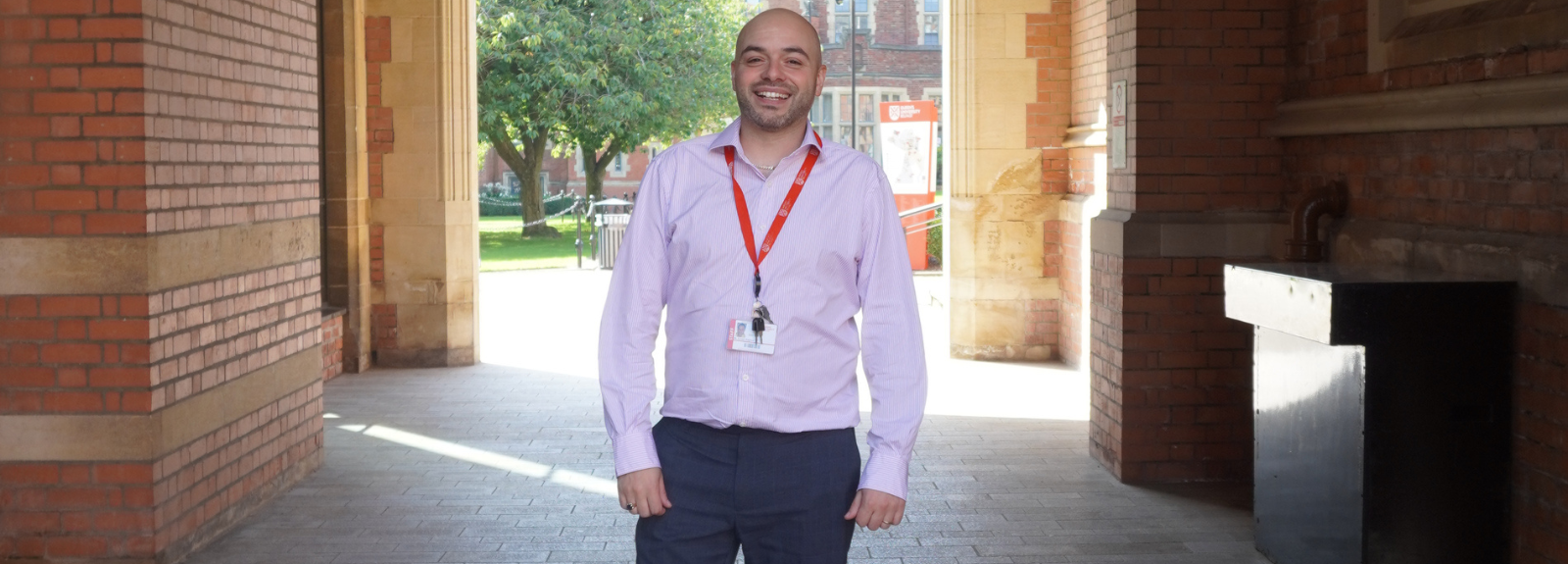 Bald smiling male standing in an arched walkway in front of a sunlit green area.  He is wearing a light pink open buttoned shirt and dark grey trousers, as well as a red lanyard around his neck.